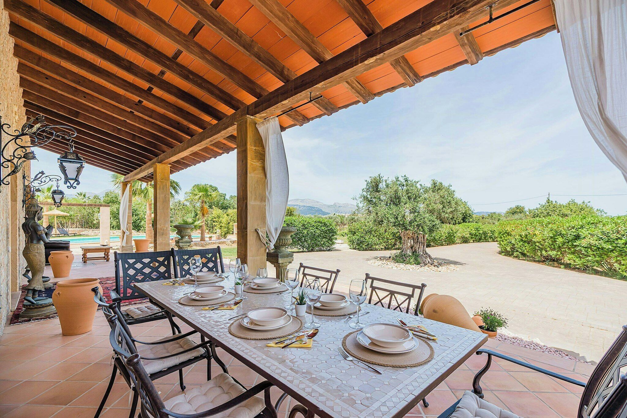 An outdoor dining area with a table set for six, surrounded by chairs, under a wooden pergola with a view of the landscape.
