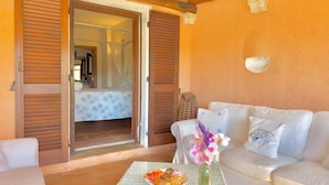 A room with a white sofa, a table with a plate of pastries, a vase with flowers, and a view of a bathroom through a door. - Villa Iole by My Villa Collection (Cala di Volpe)
