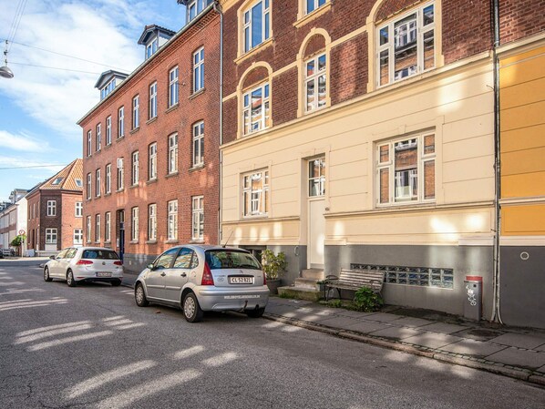 A street with parked cars and a row of buildings with multiple windows and doors.