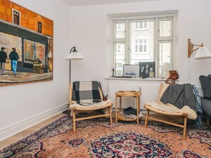 A room with a large painting, a window with blinds, a chair, a small table, and a patterned rug.
