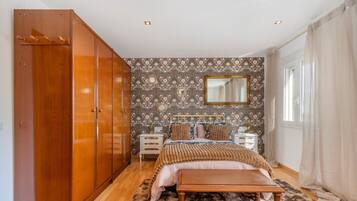 Une chambre avec une armoire en bois, un lit doté d’une tête de lit à motifs, une table de chevet, un miroir et une fenêtre avec des rideaux.