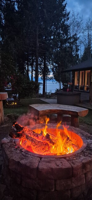 A fire pit with a stone fire ring, surrounded by a wooden deck and a view of the water. - SeaView Getaway - B.& L.T., -bed and local treats-, Retreat. (Sechelt)