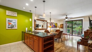 Une cuisine avec un mur vert, des armoires en bois, un îlot central en verre, des luminaires suspendus et un coin repas avec des chaises et une table.