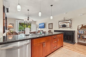A modern kitchen with a central island, stainless steel appliances, wooden cabinets, pendant lighting, and a view to the outdoors. - Wailea Blue Golf Course Condo-Walk to Shops (Kihei)