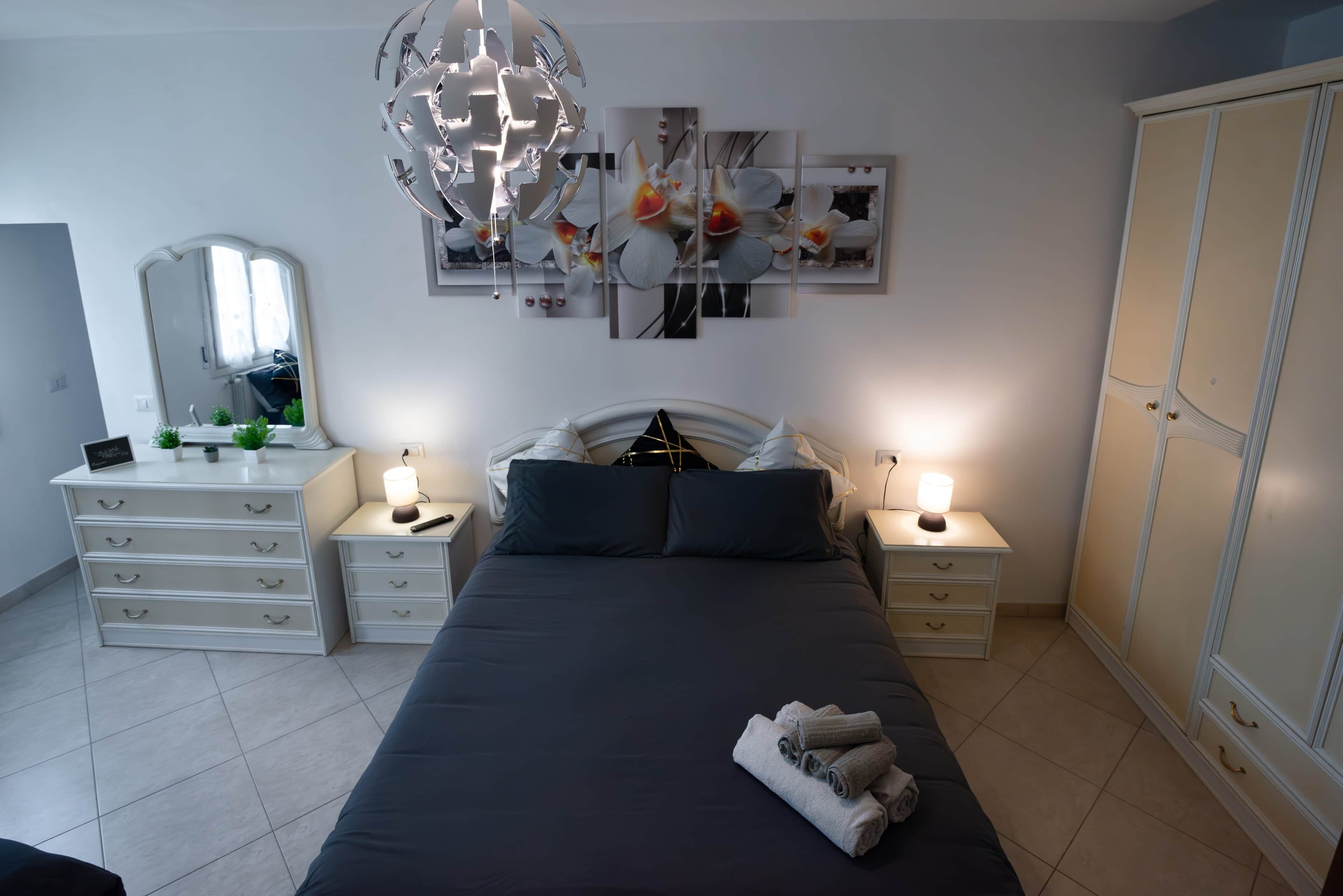 Une chambre à coucher comprenant un lit, des tables de chevet, une commode, une armoire, un lustre et des œuvres d’art murales.