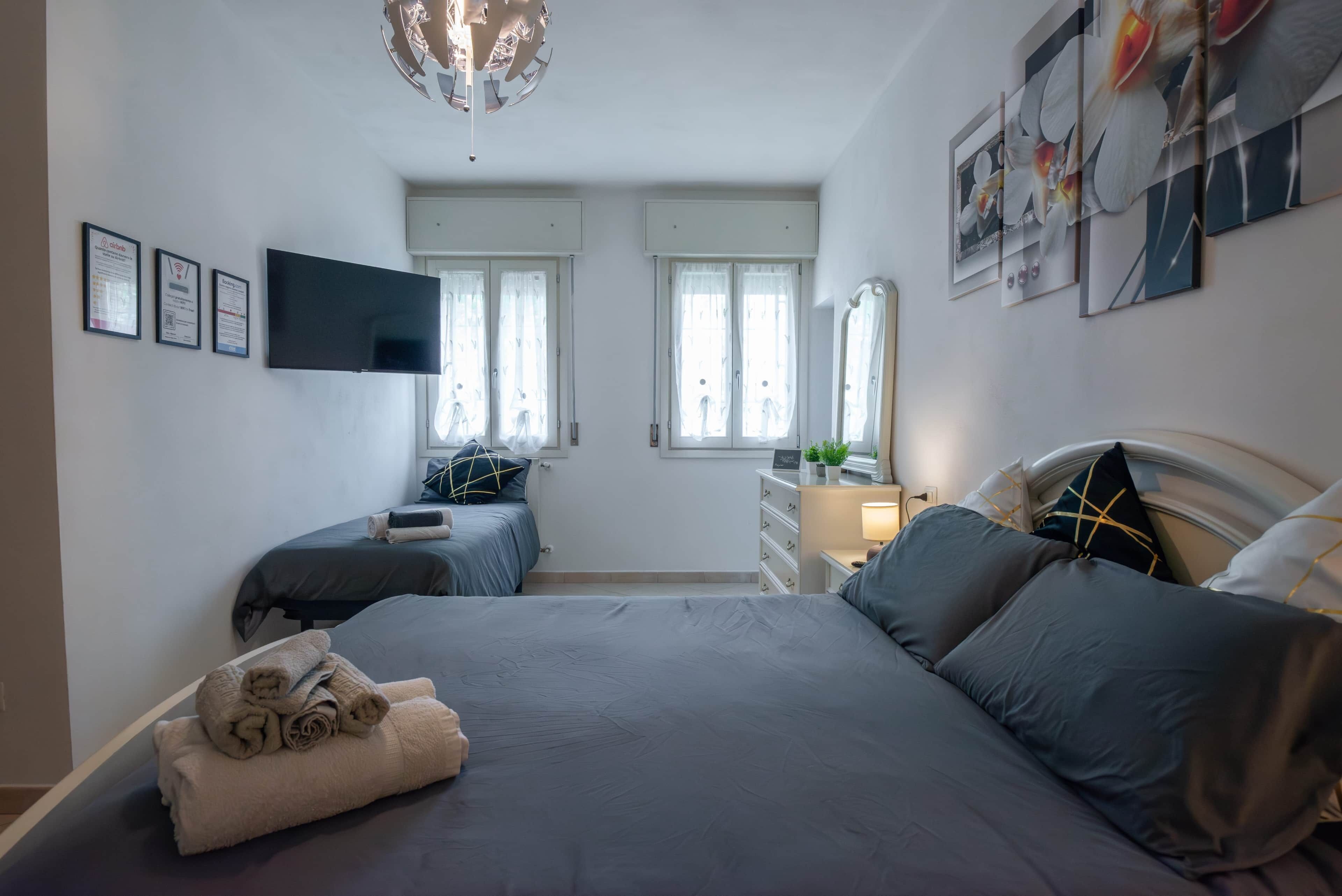A bedroom with a bed, a desk, a chair, a flat-screen TV, and a window with curtains.