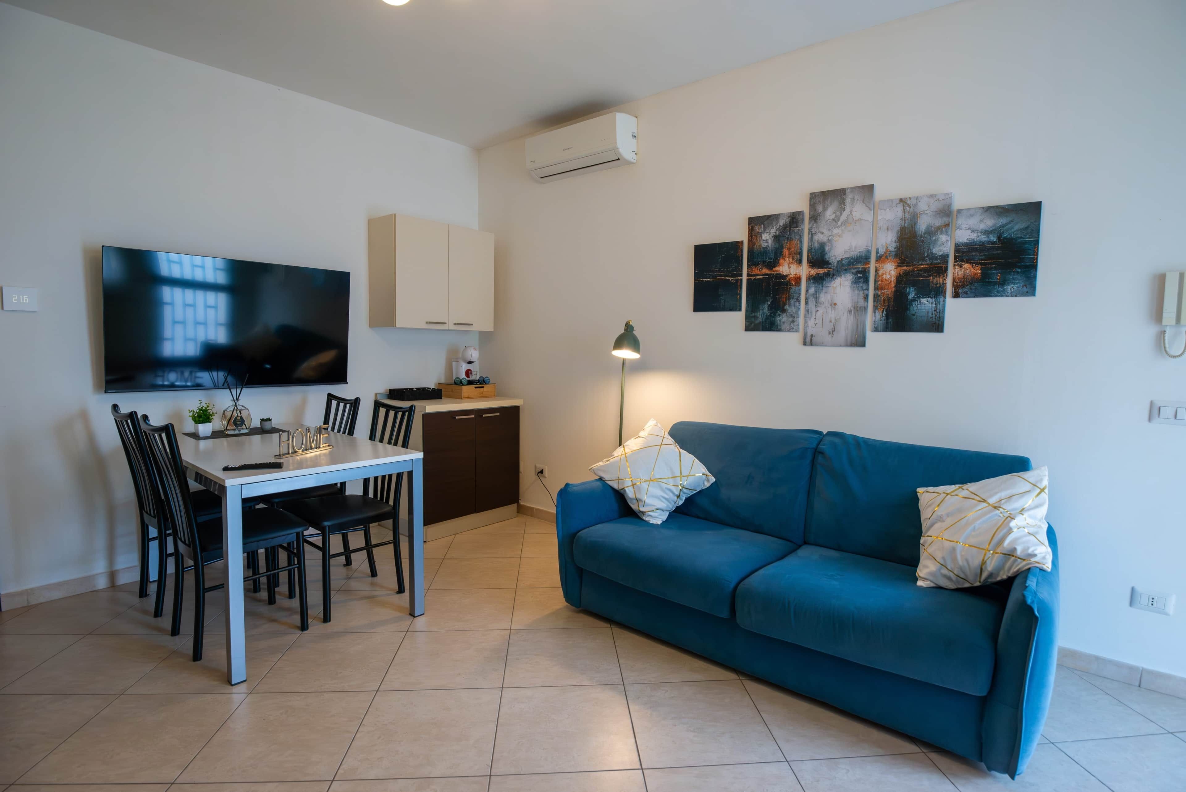 Una sala de estar moderna con un televisor de pantalla plana, una mesa de comedor con sillas, un sofá azul y arte abstracto en la pared.