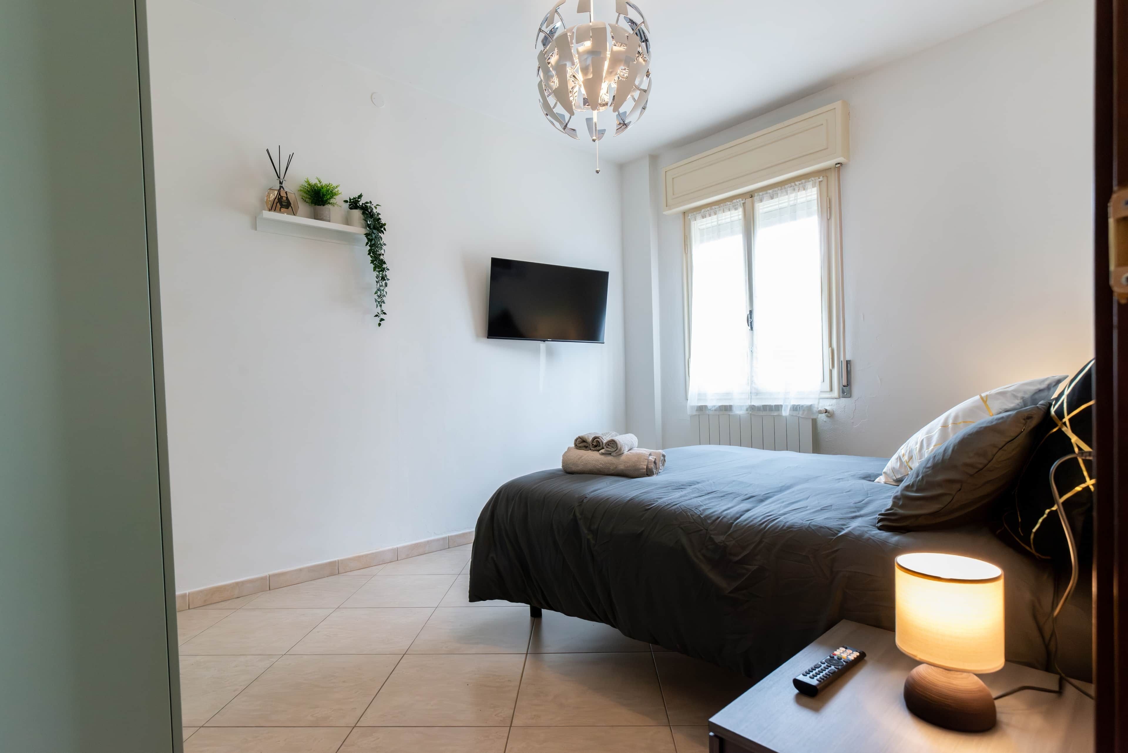Un dormitorio con una cama, un televisor montado en la pared, una lámpara de noche y una ventana con cortinas.