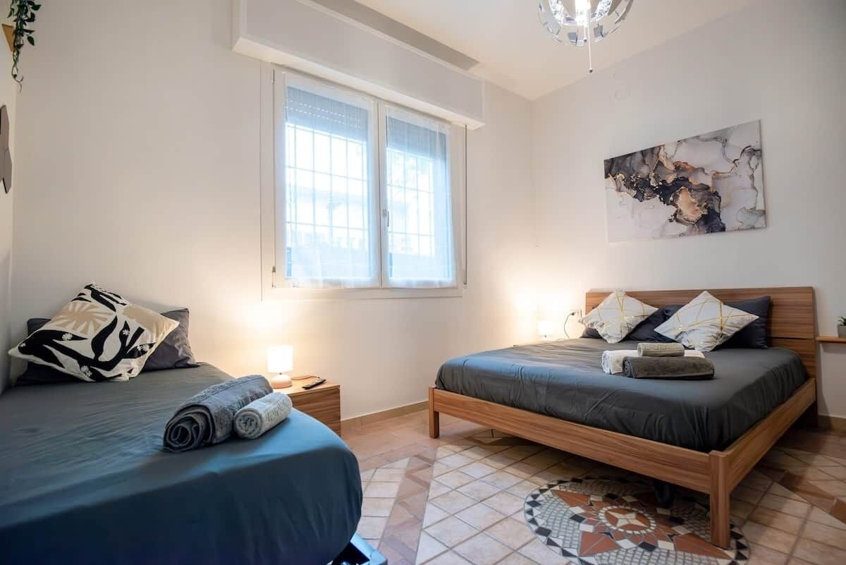 A bedroom with two beds, a window with blinds, a painting on the wall, and a patterned rug.
