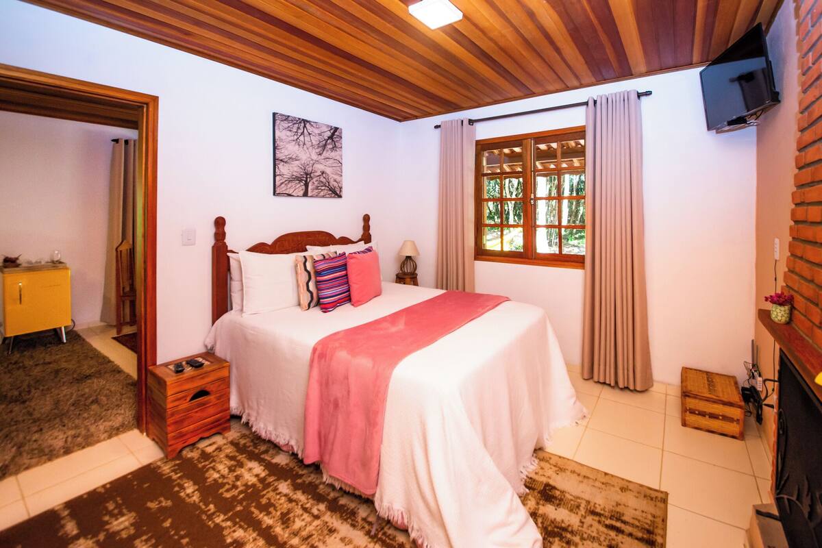 Ein Schlafzimmer mit Holzboden, einem Bett mit weißer und pinkfarbener Bettdecke, einem Fenster mit Vorhängen, einem an der Wand montierten Fernseher und einer hölzernen Kommode.