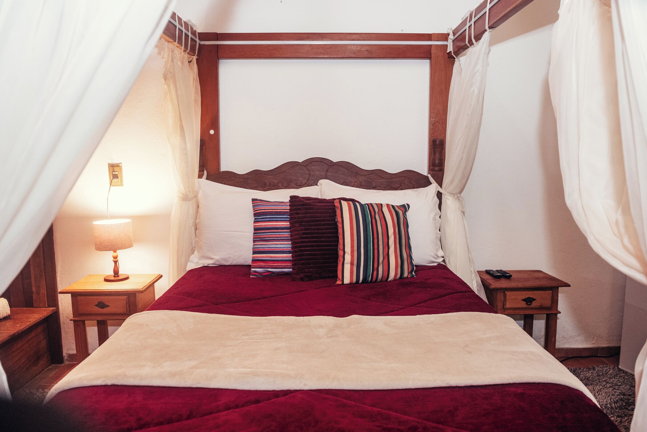 A bed with a red bedspread, multiple pillows, a wooden headboard, and a bedside table with a lamp.