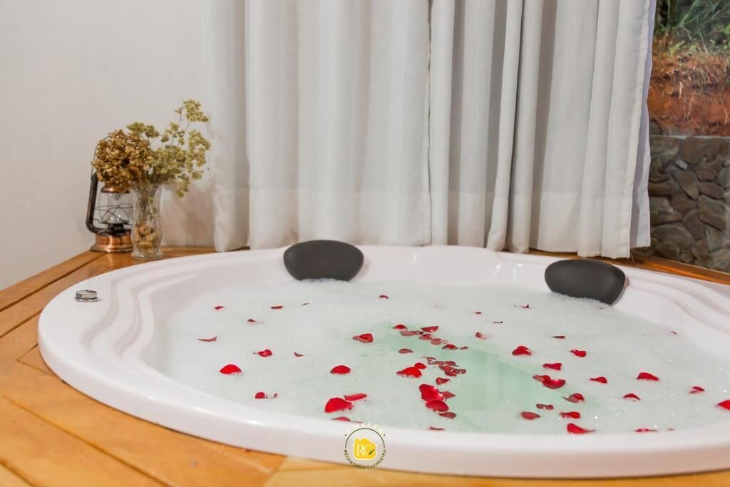 A hot tub with rose petals, a glass vase with dried flowers, and a stone wall in the background.
