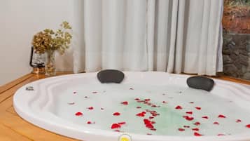 A hot tub with rose petals, a glass vase with dried flowers, and a stone wall in the background.