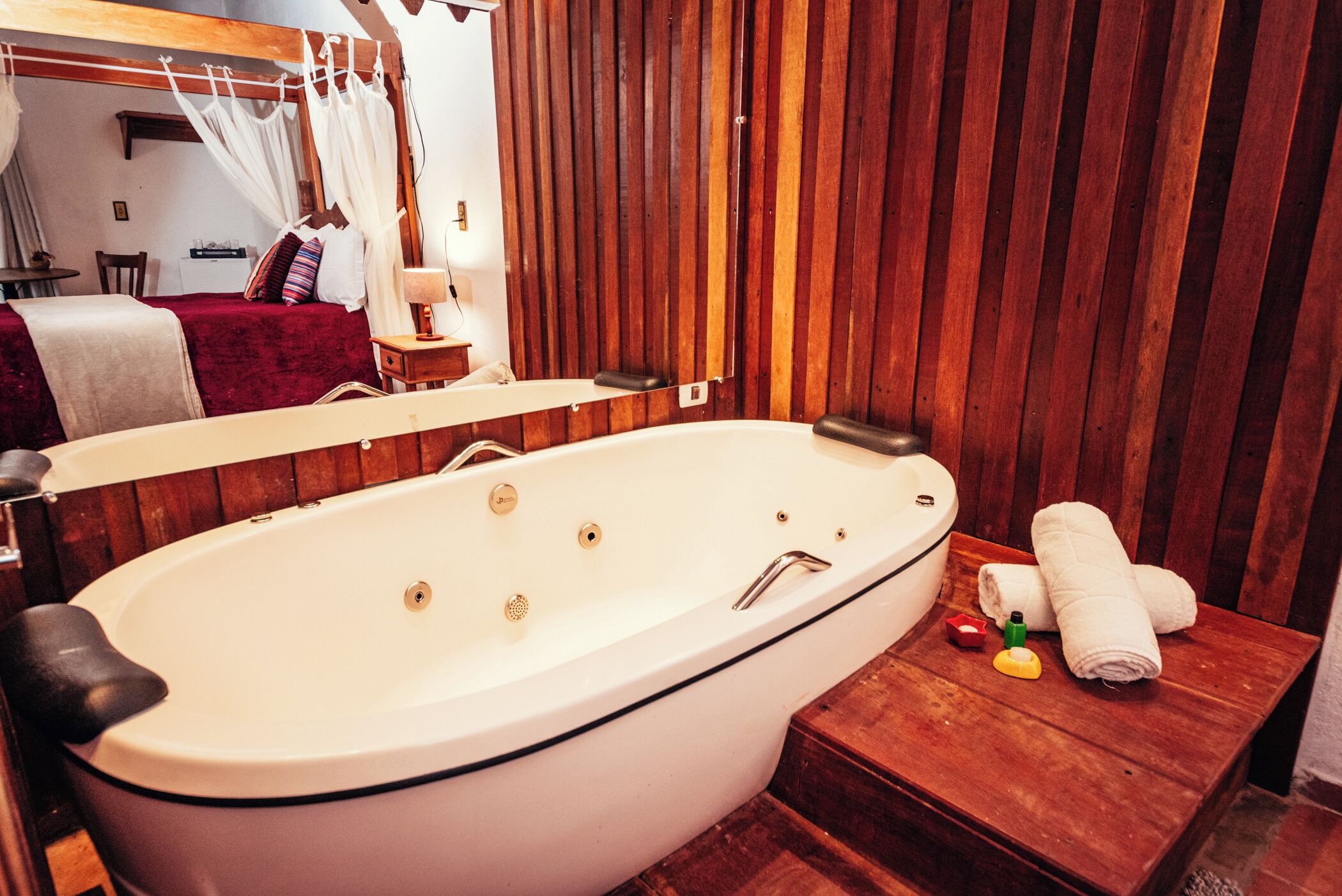 A bathroom with a large bathtub, wooden paneling, a vanity with a mirror, and a bedside table with towels.