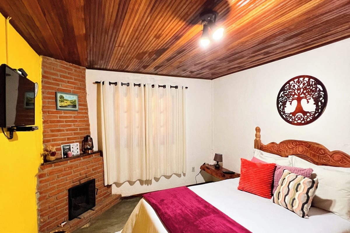 Ein Schlafzimmer mit Kamin aus Backsteinen, einem Bett mit roter Decke, Holzboden und einem an der Wand befestigten Fernseher.