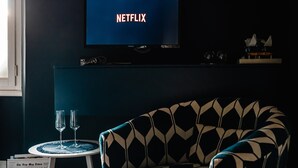 A room with a television displaying 'NETFLIX,' a patterned armchair, a small table with two glasses, and a newspaper. - 1890 – Deluxe Room – Whirlpool Bath – Wi-Fi – Air Conditioning (Chiavari)