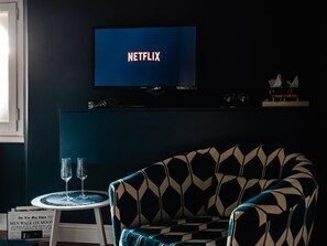 A room with a television displaying 'NETFLIX,' a patterned armchair, a small table with two glasses, and a newspaper. - 1890 – Deluxe Room – Whirlpool Bath – Wi-Fi – Air Conditioning (Chiavari)