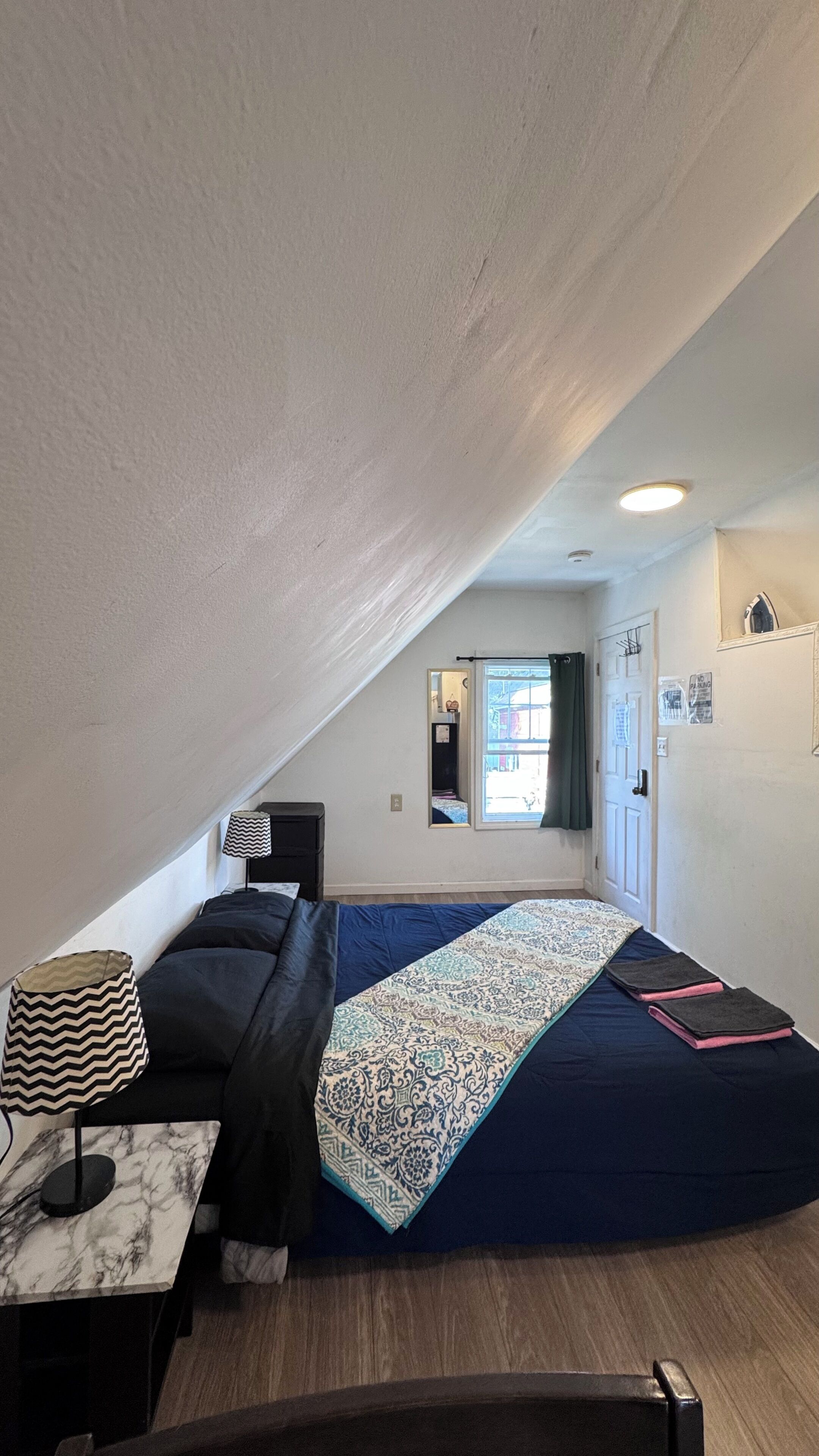 A small, slanted room with a bed, a desk, a chair, a lamp, and a window with curtains.