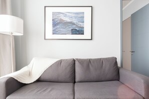 A grey sofa with a white throw blanket, a framed picture of ocean waves, a floor lamp, and a door in the background. - Departamento 4 Pax Barrio Italia (Santiago)