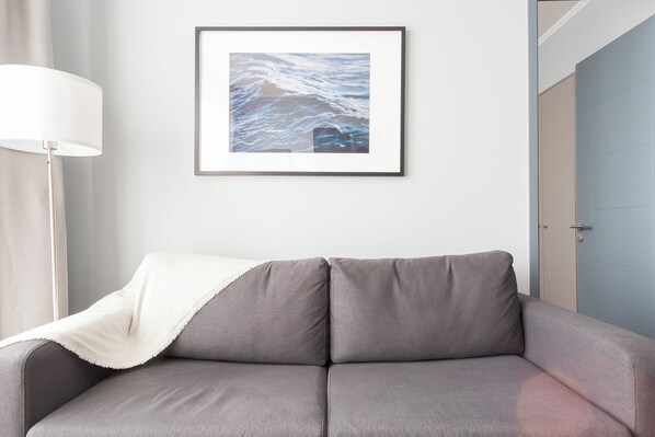 A grey sofa with a white throw blanket, a framed picture of ocean waves, a floor lamp, and a door in the background. - Departamento 4 Pax Barrio Italia (Santiago)