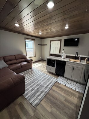 Una sala de estar moderna con un sofá de cuero marrón, una zona de cocina con encimera y armarios, y techo de madera.