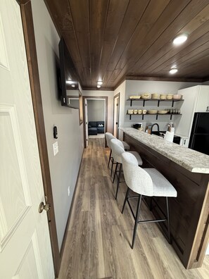 Eine moderne Küche mit Holzbalkendecke, einer integrierten Bar mit Barhocker und einem sichtbaren Wohnbereich mit einer Couch.