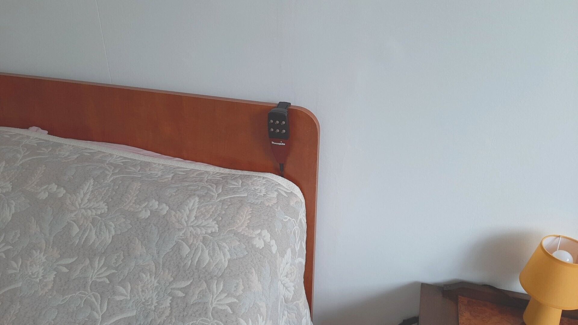 A bed with a wooden headboard, a remote control on it, and a yellow lamp on a nightstand.
