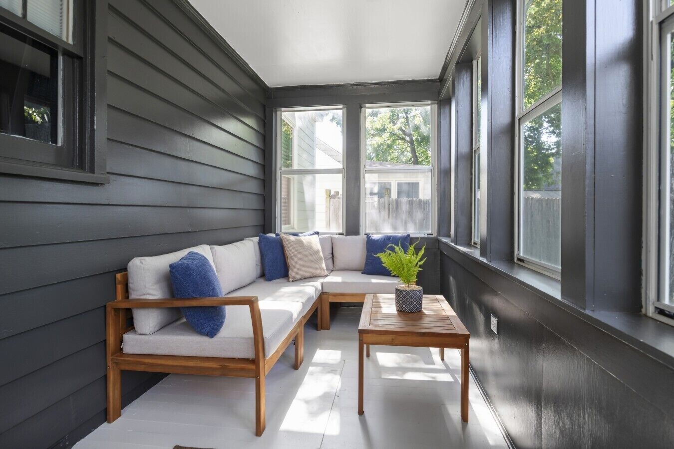 A covered porch with a wooden bench and a table, a gray sofa with blue cushions, and a potted plant.