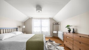 Une chambre à coucher comprenant un lit, une table de chevet, une lampe, un radiateur, une commode en bois et une fenêtre avec des rideaux.