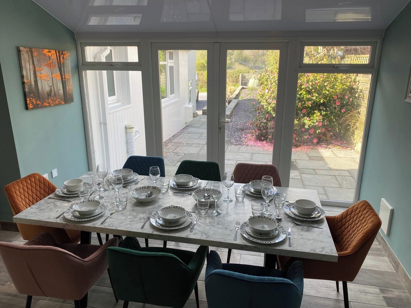 A dining area with a table set for breakfast, surrounded by chairs, and a view of a garden through large windows.