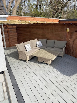 A covered outdoor seating area with a wicker sofa and cushions, a wooden deck, and a wooden wall with a warning sign. - Spacious Family Getaway Hot Tub, Sauna & Garden (Conwy Principal Area)