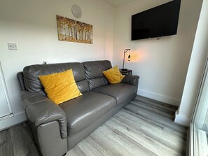 Ein modernes Wohnzimmer mit einem Ledersofa, zwei senfgelben Kissen, einem an der Wand befestigten Flachbildfernseher und einer Stehlampe.
