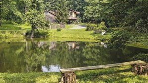 A serene lakeside setting with a wooden bench, a rustic house, and lush greenery. - Hudson Valley Private Retreat with 3 bedrooms, 2 living spaces, and a pond. (Hillsdale)