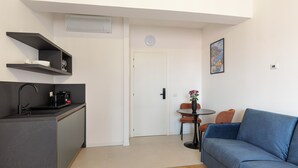 A modern kitchen with a black countertop, a built-in oven, and a coffee machine. There is a dining area with a blue sofa and a small round table. - Pharmacy B (stresa)