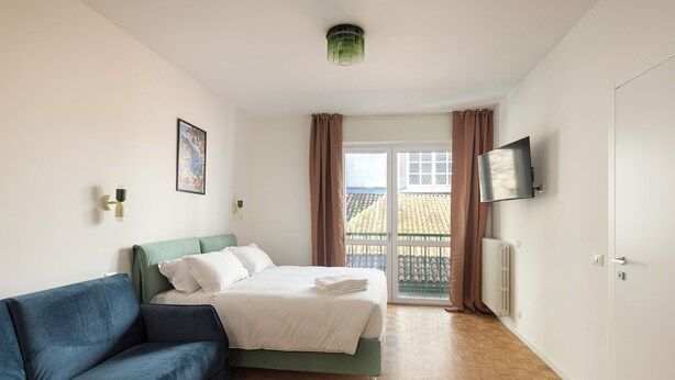 A bedroom with a bed, a blue sofa, a TV on the wall, and a view of the outside. - Pharmacy B (stresa)