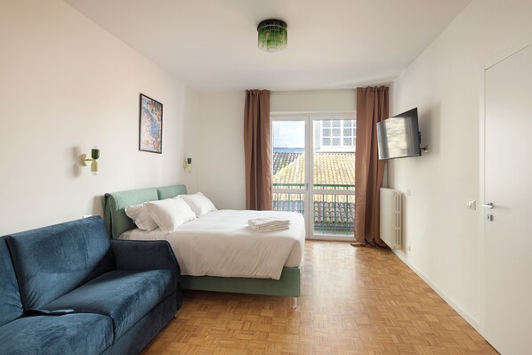 A bedroom with a bed, a blue sofa, a TV on the wall, and a view of the outside. - Pharmacy B (stresa)
