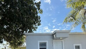 A two-story house with a white exterior, a front door, two windows, and a small porch area with three chairs and a potted plant. - Cozy Palm Retreat 2BR Home • Near Beaches (Lake Worth Beach)