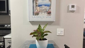 A dining area with a table, chairs, a potted plant, a framed picture, and a thermostat on the wall. - Cozy Palm Retreat 2BR Home • Near Beaches (Lake Worth Beach)