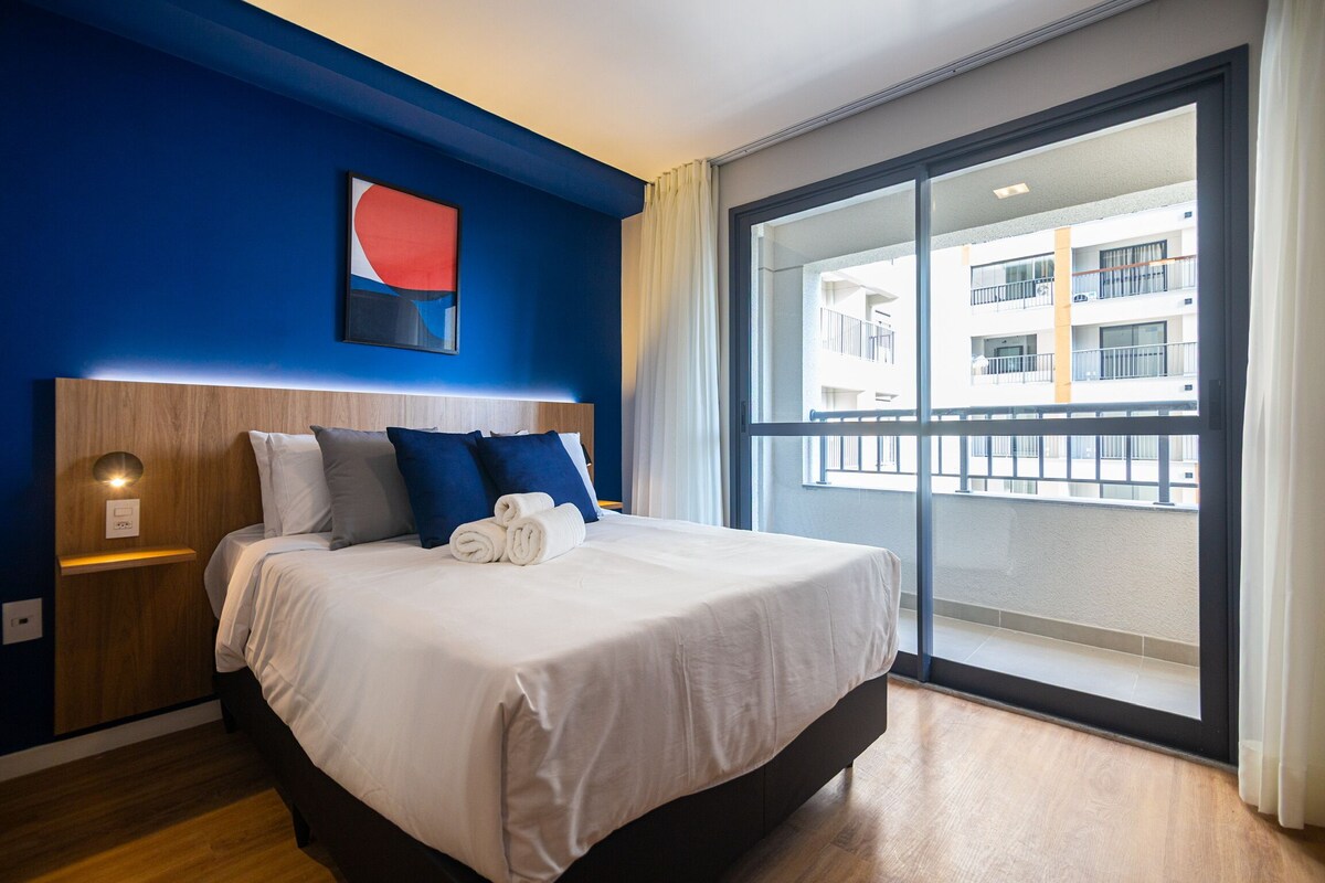 A hotel room with a large bed, a nightstand, a wall-mounted light, and a view of a building through a large window.