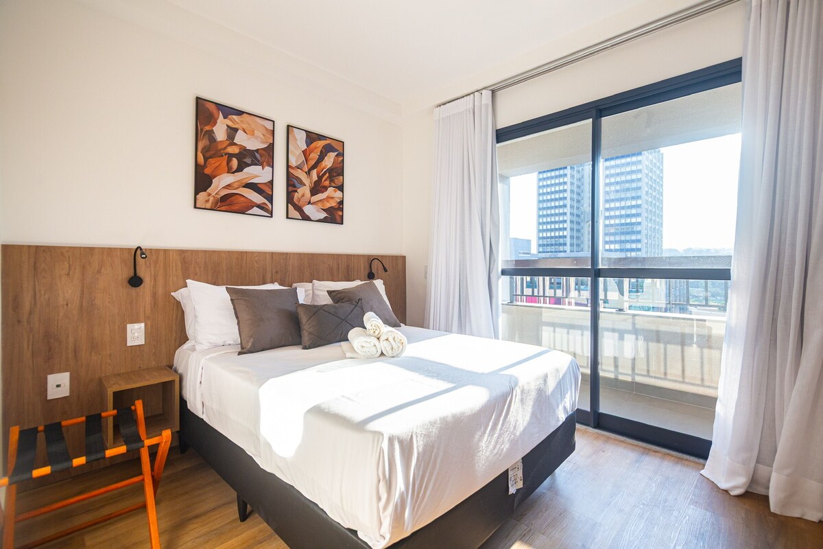 A modern hotel room with a large bed, a wooden headboard, two wall-mounted artworks, a small wooden nightstand, and a view of the city through a large window.
