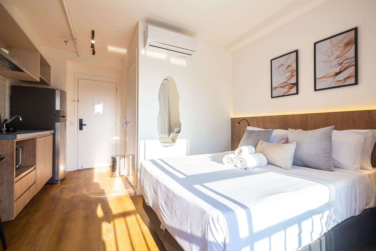 A hotel room with a bed, a desk, a mirror, and a wall-mounted air conditioner.