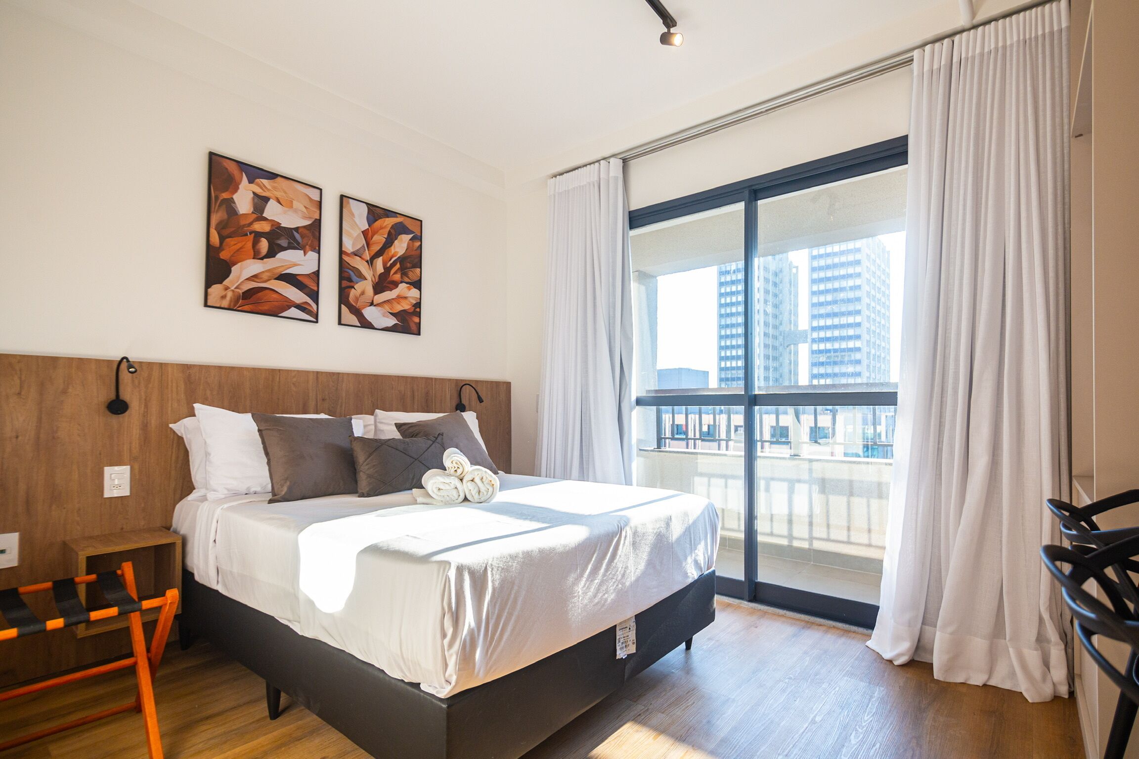 Habitación de hotel con una cama grande, cabecera de madera, dos cuadros abstractos, vista a la ciudad a través de un ventanal amplio y un balcón con una silla.