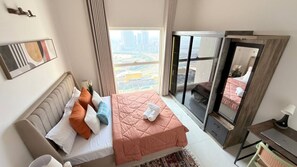 A hotel room with a bed, a desk, a chair, a window with a city view, and a sliding door. - Beige Zenith : 2BHK Apt 32nd floor + Pool (Gandhinagar)