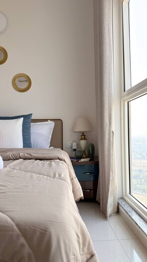 A bedroom with a bed, bedside table, lamp, and a view through a window. - Beige Zenith : 2BHK Apt 32nd floor + Pool (Gandhinagar)