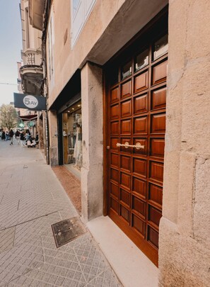 A building with a large wooden door, a sign above the door, and a sidewalk with people walking. - Go to Downtown Pontevedra (Pontevedra)