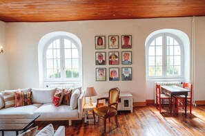 Une pièce avec un plancher en bois, des murs blancs, un canapé, une table et deux chaises. Il y a deux fenêtres et un radiateur.