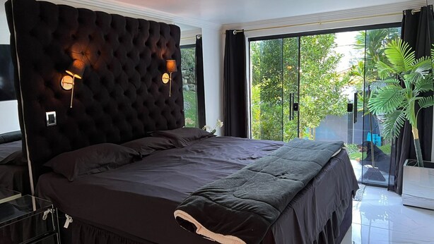Un dormitorio con cabecera capitonada, cama con ropa de cama oscura, mesita de noche, lámpara y vistas al exterior con vegetación.