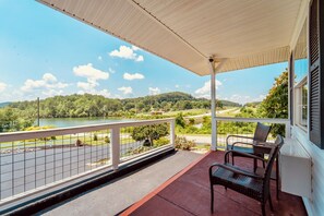 A covered balcony with a view of a lake and hills, furnished with two chairs and a table.