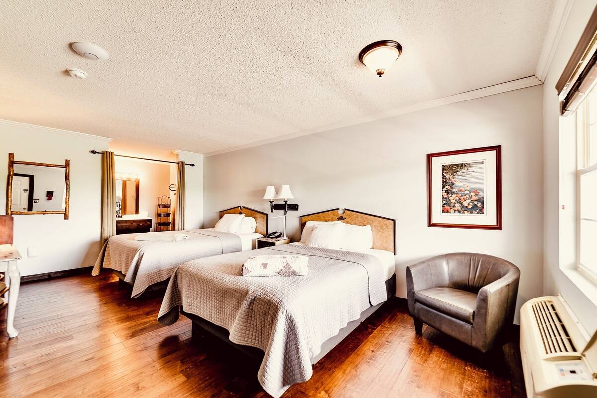 A hotel room with two beds, a chair, a desk, a mirror, a picture on the wall, and a window.