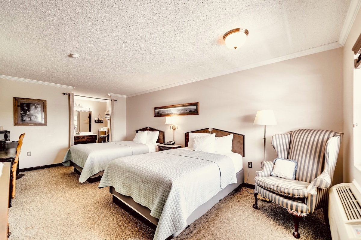 A hotel room with two beds, a chair, a desk, a mirror, and a lamp.